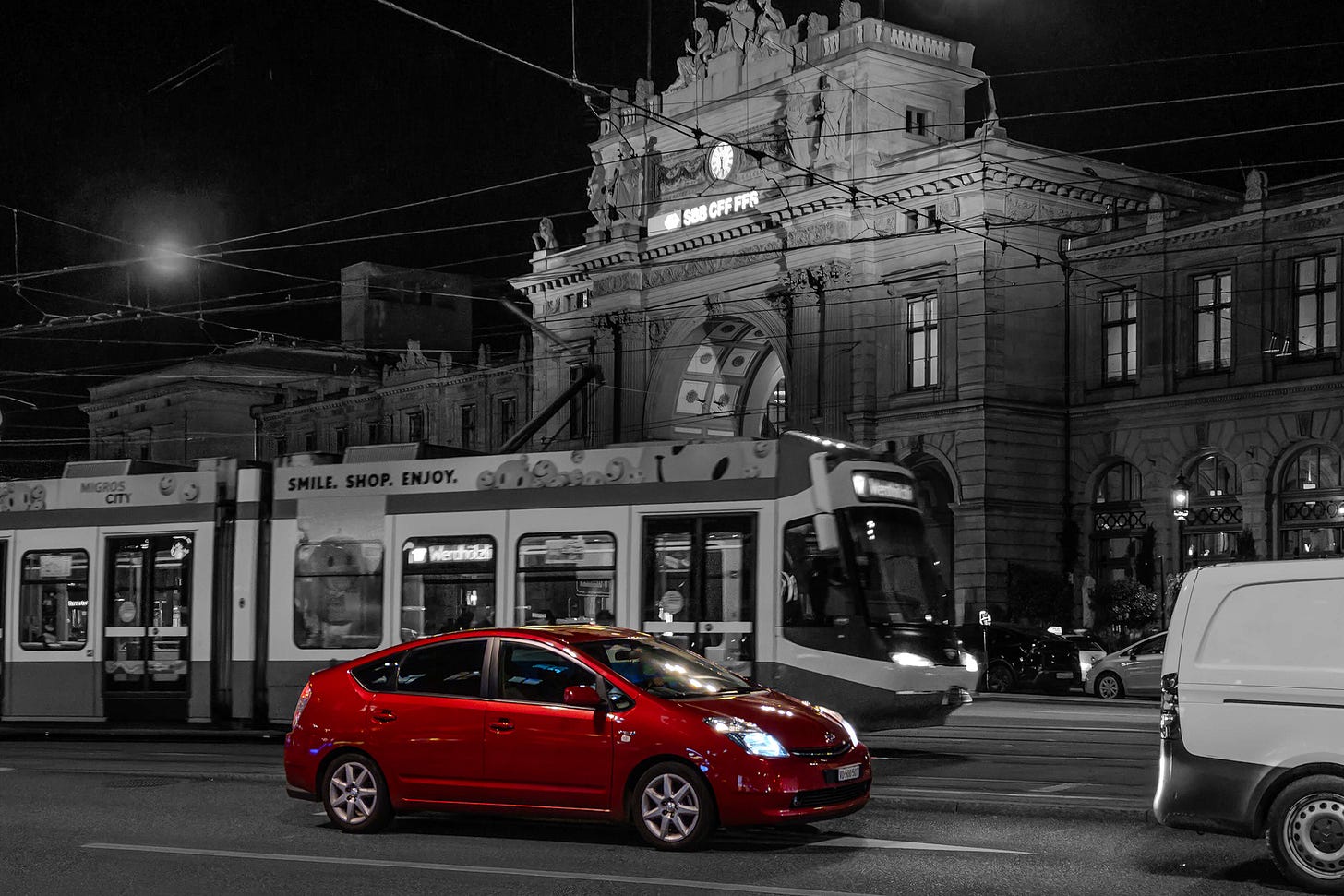 Ein rotes Auto fährt über den Zürcher Bahnhofplatz, während eine Tram vorbeifährt. Das Foto ist schwarz-weiss gehalten, nur das Auto ist farbig hervorgehoben. Oberhalb des Bahnhofportals leuchtet das SBB-Logo und eine Uhr zeigt die Zeit an. Ein rotes Auto fährt über den Zürcher Bahnhofplatz, während eine Tram vorbeifährt. Das Foto ist schwarz-weiss gehalten, nur das Auto ist farbig hervorgehoben. Oberhalb des Bahnhofportals leuchtet das SBB-Logo und eine Uhr zeigt die Zeit an.