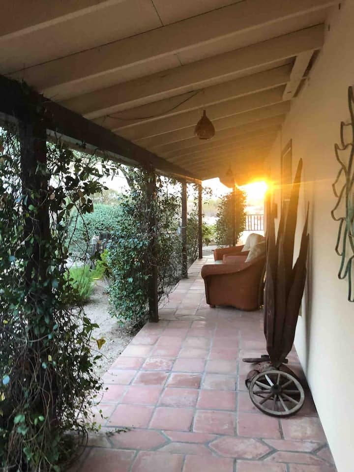 A peaceful covered porch at sunset with warm golden light streaming in, surrounded by greenery, inviting rest and reflection—like a space prepared in advance, waiting to be enjoyed.