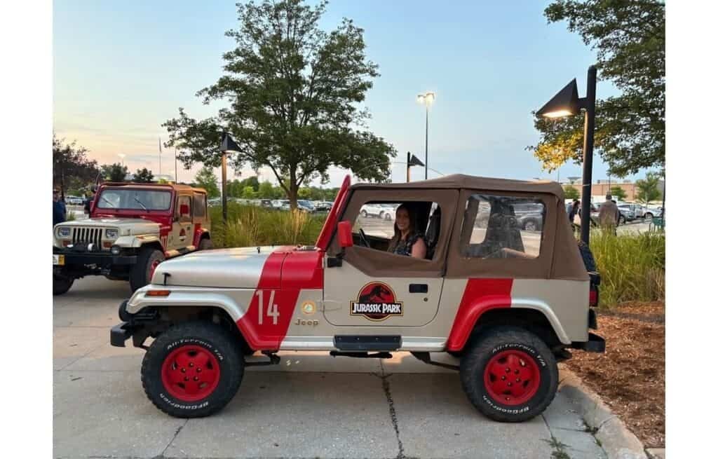 A woman sits in a Jurassic Park-themed Jeep Wrangler with a tan soft top and red accents, parked outdoors amid trees and another Jeep—ready for cake or Connor’s Big News, no sign of chaos in this adventurous scene.