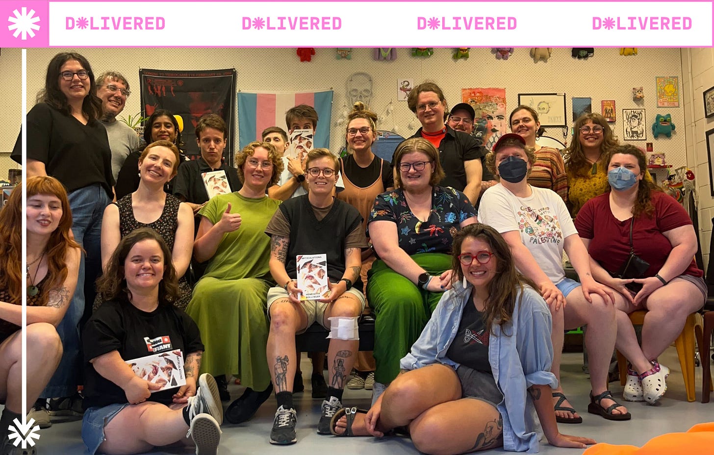 About 20 people gathered in a room in front of a decorated wall. People are standing and sitting on chairs and on the floor and smiling at the camera