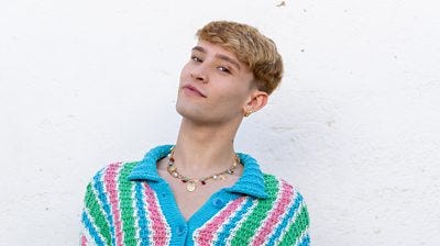 Jack S wears a bright knitted cardigan with green blue and pink stripes. It is unbuttoned at the top to show a necklace with gold shells, pearls and colourful beads