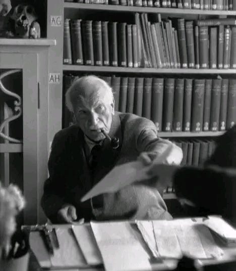 Black-and-white photograph of Carl Jung seated at his desk in his home library in Küsnacht, Switzerland. He is holding out a piece of paper, with shelves of books behind him.