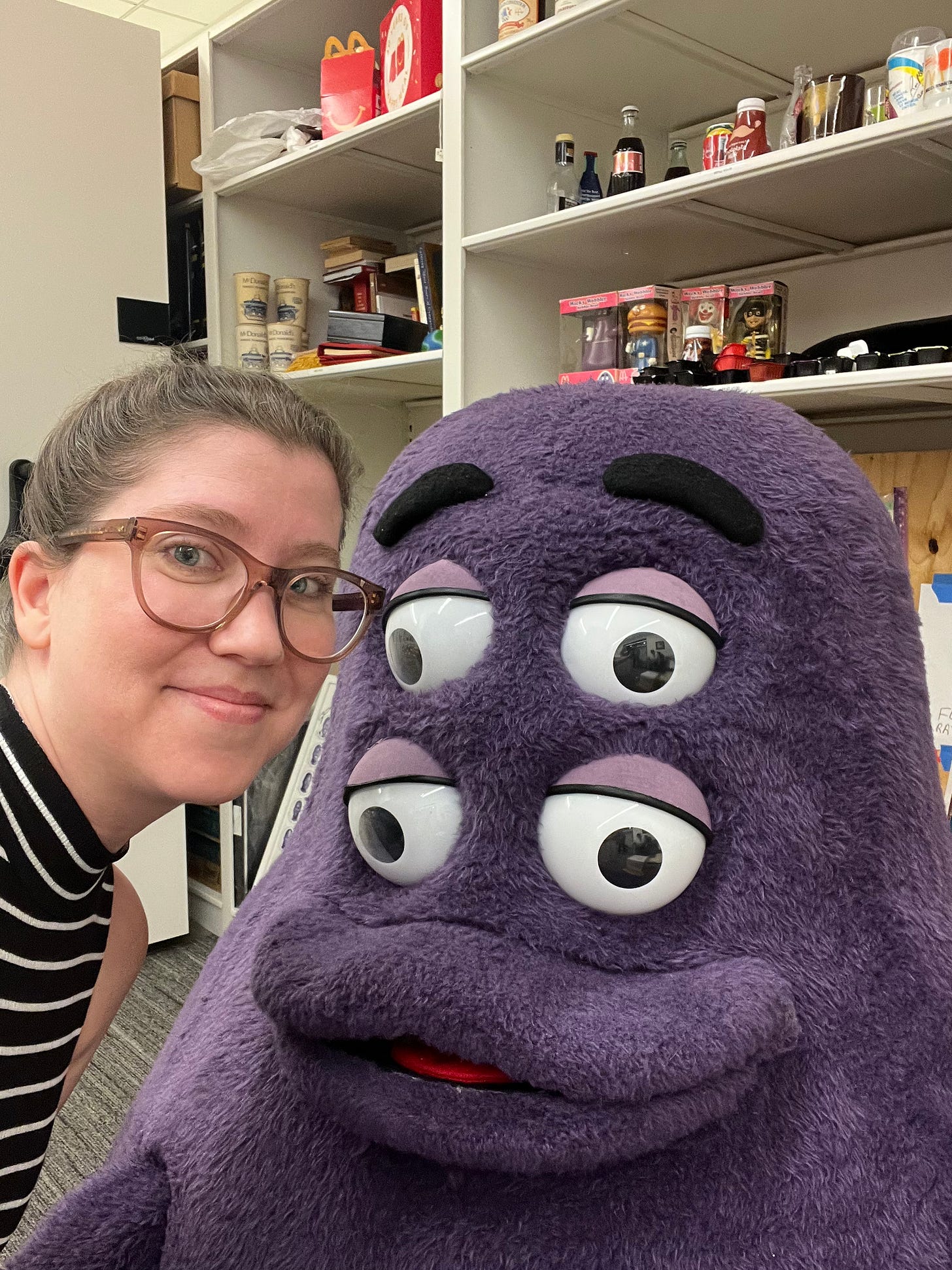 A woman with glasses leaning in to frame next to the head of a Grimace mascot with two sets of eyes; behind them are shelves filled with McDonald's memorabilia