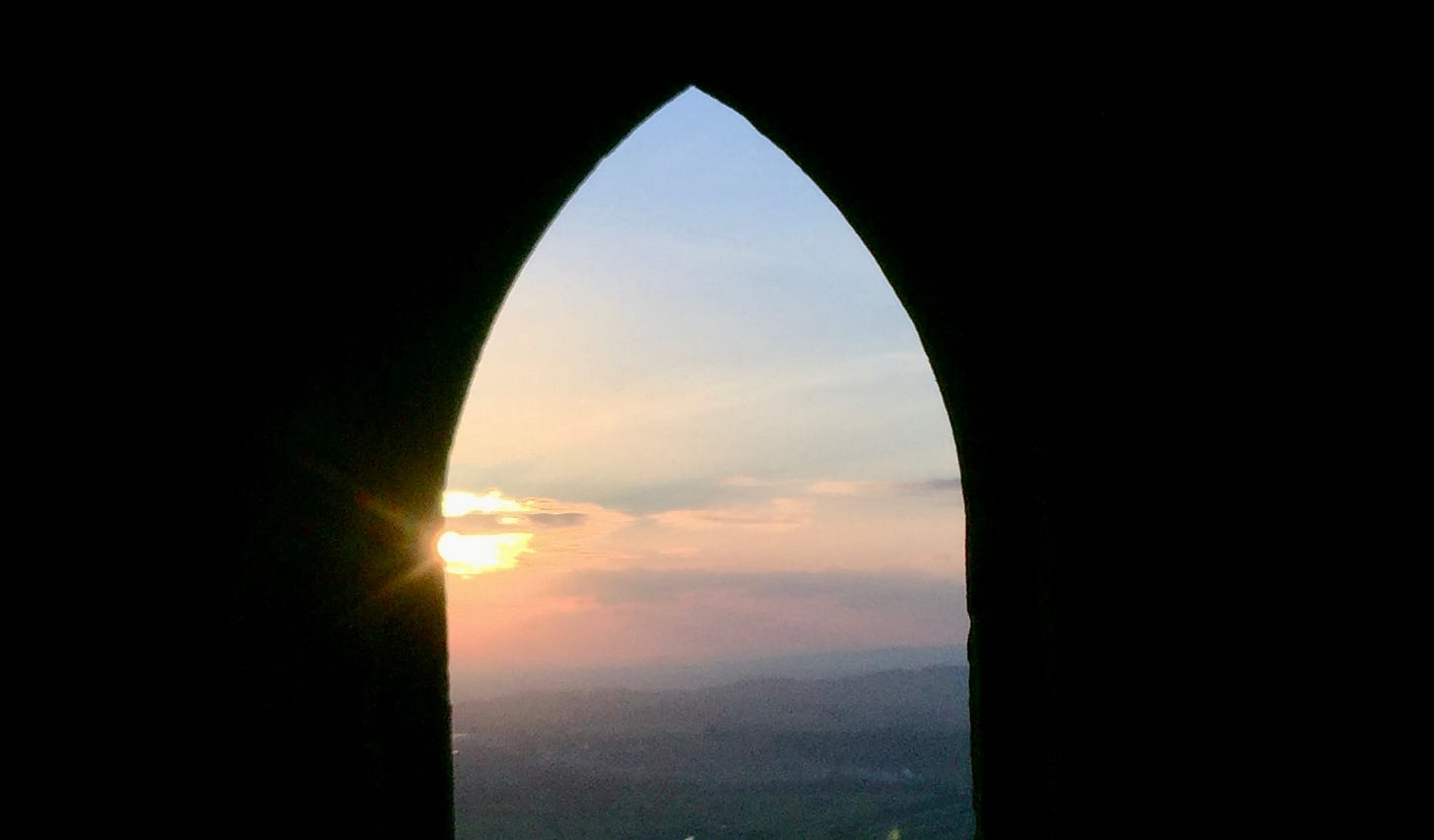 Sunrise viewed through the stone archway of St Michael's Mount, with warm light breaking over the distant landscape beyond. Sunrise viewed through the stone archway of St Michael's Mount, with warm light breaking over the distant landscape beyond.