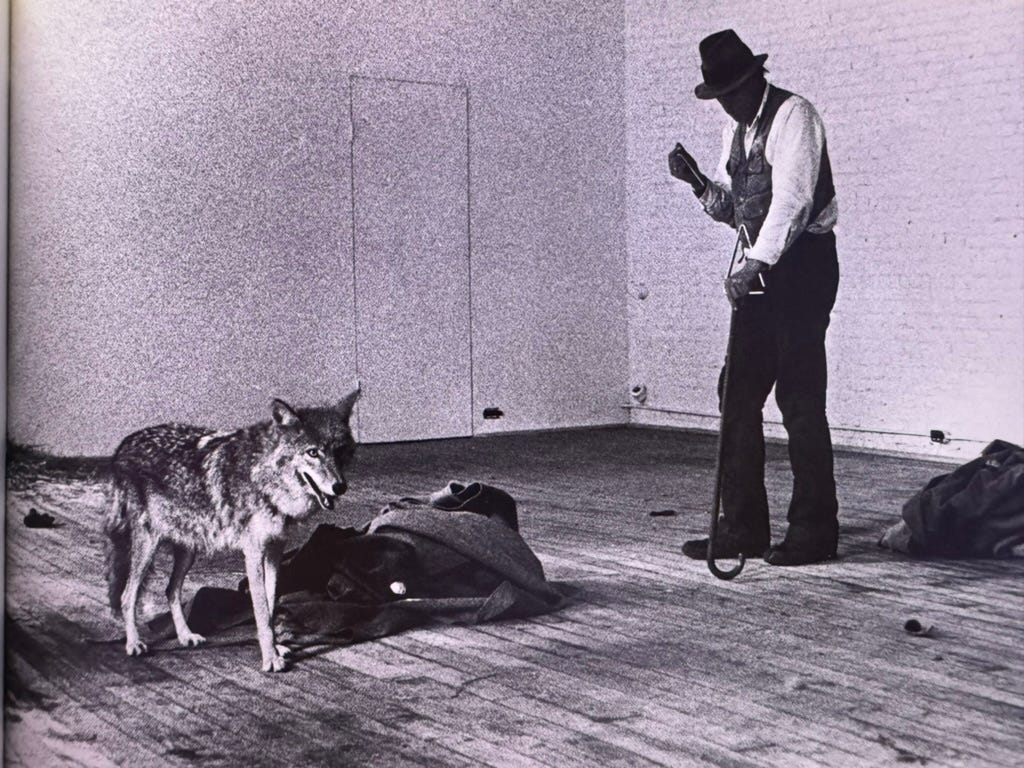 A photograph of a man and a coyote. The coyote is standing next to a bundle of rags, looking off-camera with his ears up and his eyes seeming alert. The man is standing off to his side, looking towards the coyote. There is a triangle around his neck, which he is playing with one hand; the other is on the hand of a shepherd's crook. He is wearing a white shirt, a dark vest, and a trilby.
