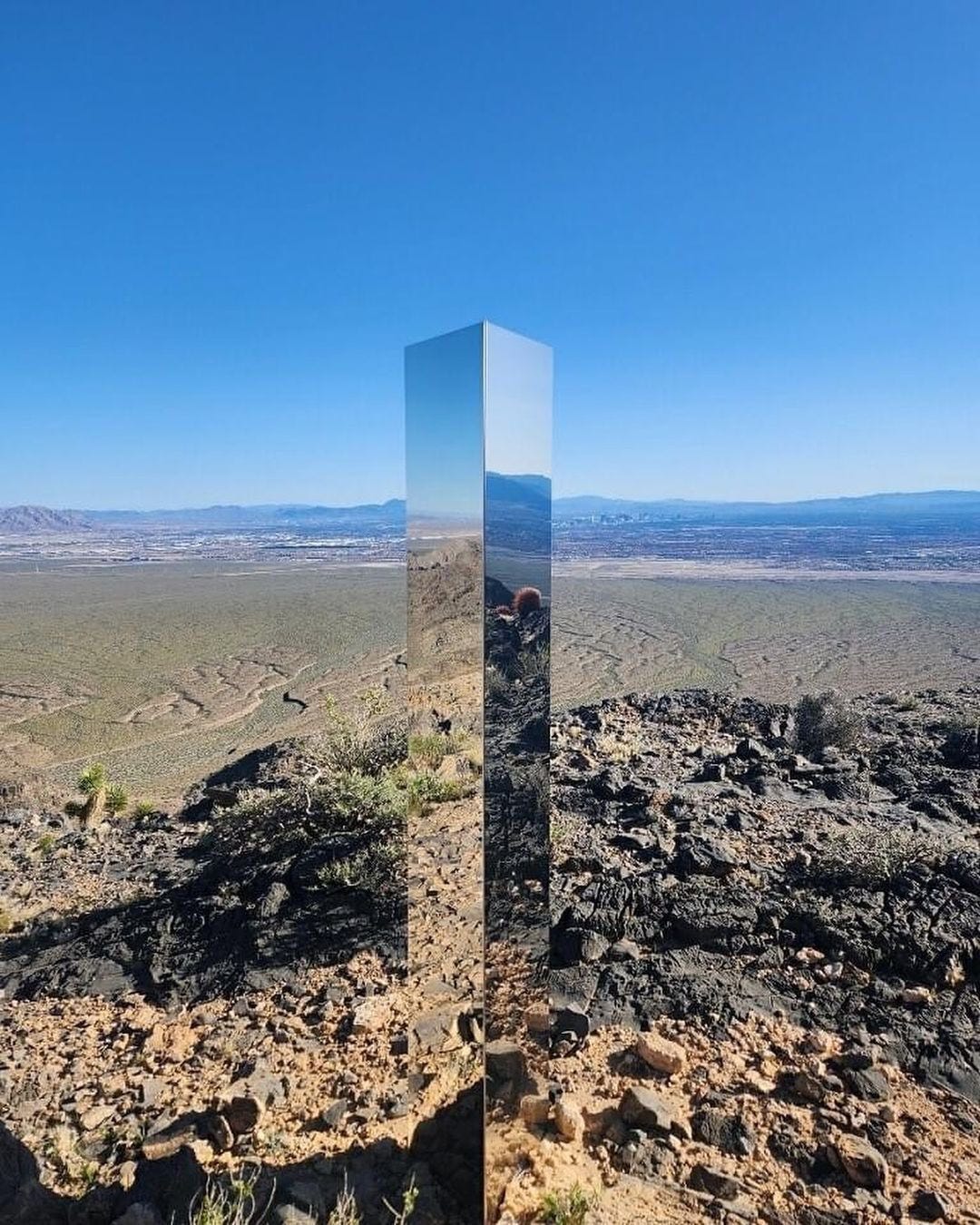 A photo of the Utah monolith, a mysterious metal three metre tall pillar that suddenly appeared in a desert canyon in 2016 until it was removed in 2020. It is a sunny day with a clear blue sky, and the shiny pillar reflects the scenery and sky surrounding it.