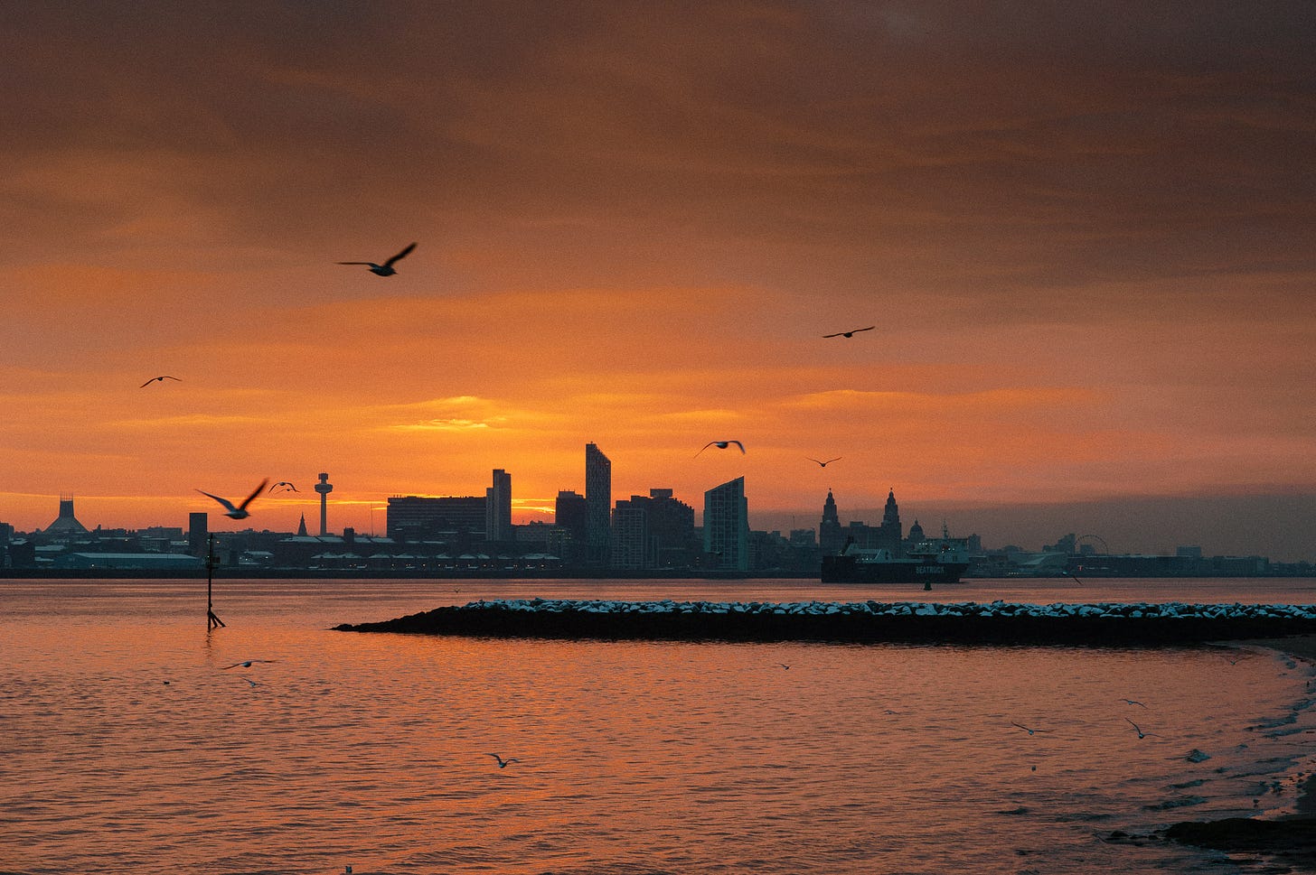 During a deep red sunrise a ship heads out the River Mersey while birds fly. There is a light dusting of snow on the rocks.
