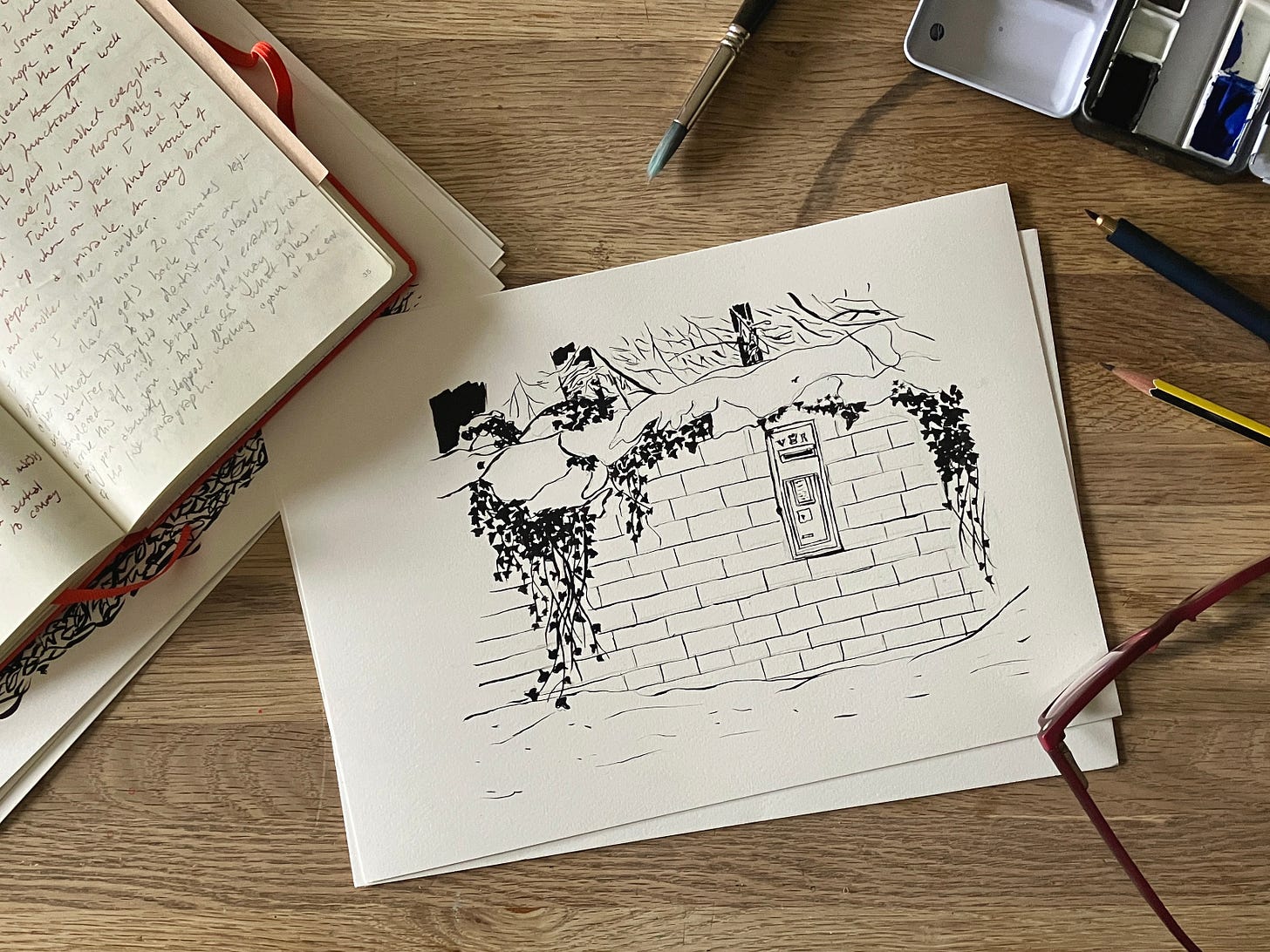 painting of snow topped wall with ivy on it and a letterbox in it sits atop a table surrounded by journals, painting and writing things