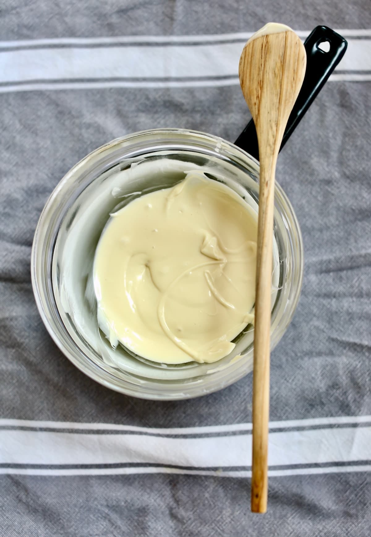 a bowl of melted white chocolate and a wooden spoon on a table with a gray tablecloth.