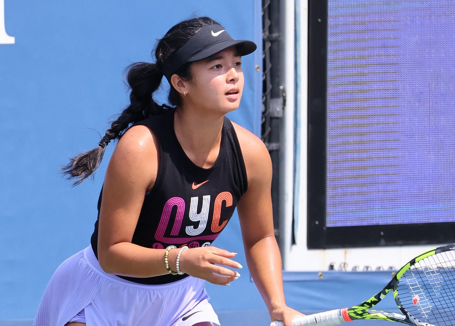 Filipina tennis player Alexandra Eala prepares to return a shot during a match on a hard court at the 2024 U.S. Open. She wears a black “NYC” tank top, white skirt, and pink sneakers, showcasing her focus and athletic form in action.
