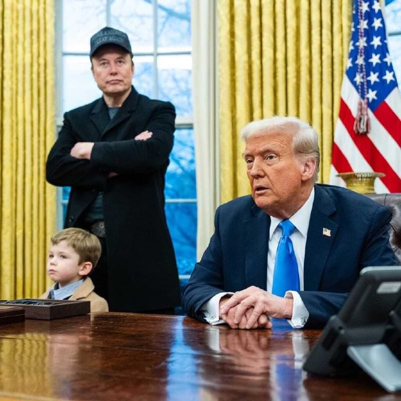 Elon Musk stands with his young son in the Oval Office, arms crossed, wearing a 'Make America Great Again' hat, while Donald Trump sits at the Resolute Desk, hands clasped. The child, dressed in a tan coat, looks off to the side, adding a lighthearted contrast to the formal setting.