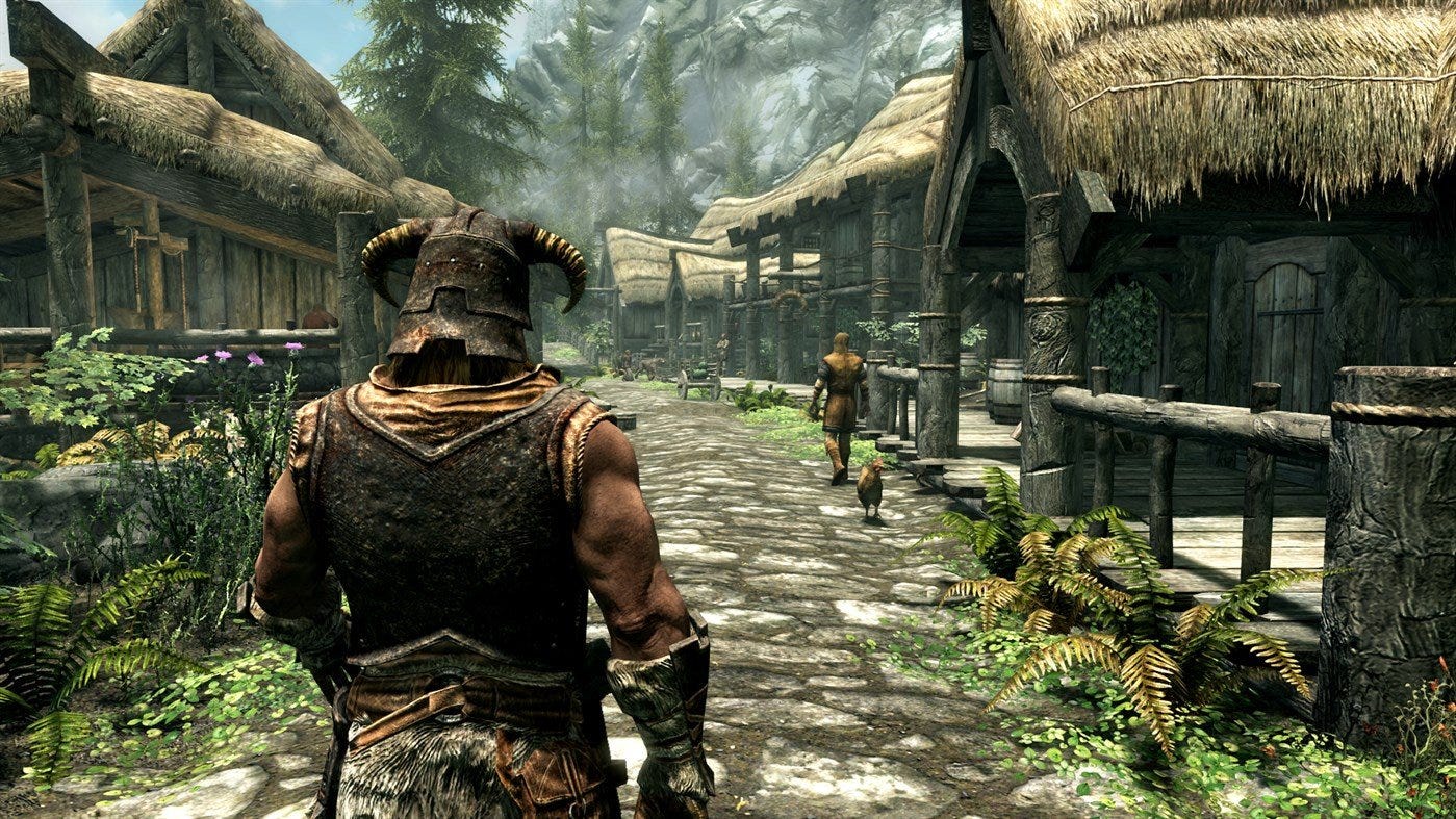 A screenshot from the video game The Elder Scrolls V: Skyrim. The image shows a snowy mountain landscape with a clear blue sky. The main character is a warrior in the foreground, wearing a steel helmet and a fur cloak. The character is facing away from the camera and is walking down a dirt path. There are other characters in the background, some of whom are villagers and others are guards. The image is from the marketing material for Skyrim, an open-world fantasy RPG by Bethesda.