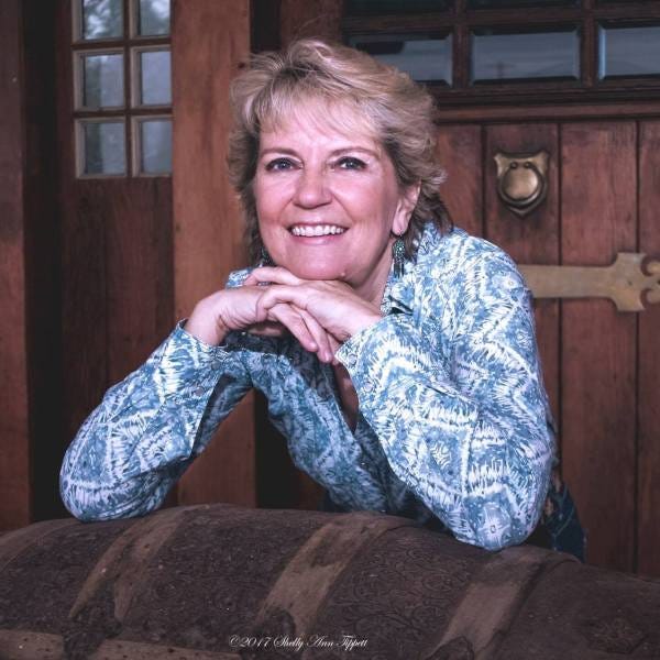 Portrait of a smiling woman leaning on an ornate barrel, wearing a light blue patterned shirt, set against a wooden background.