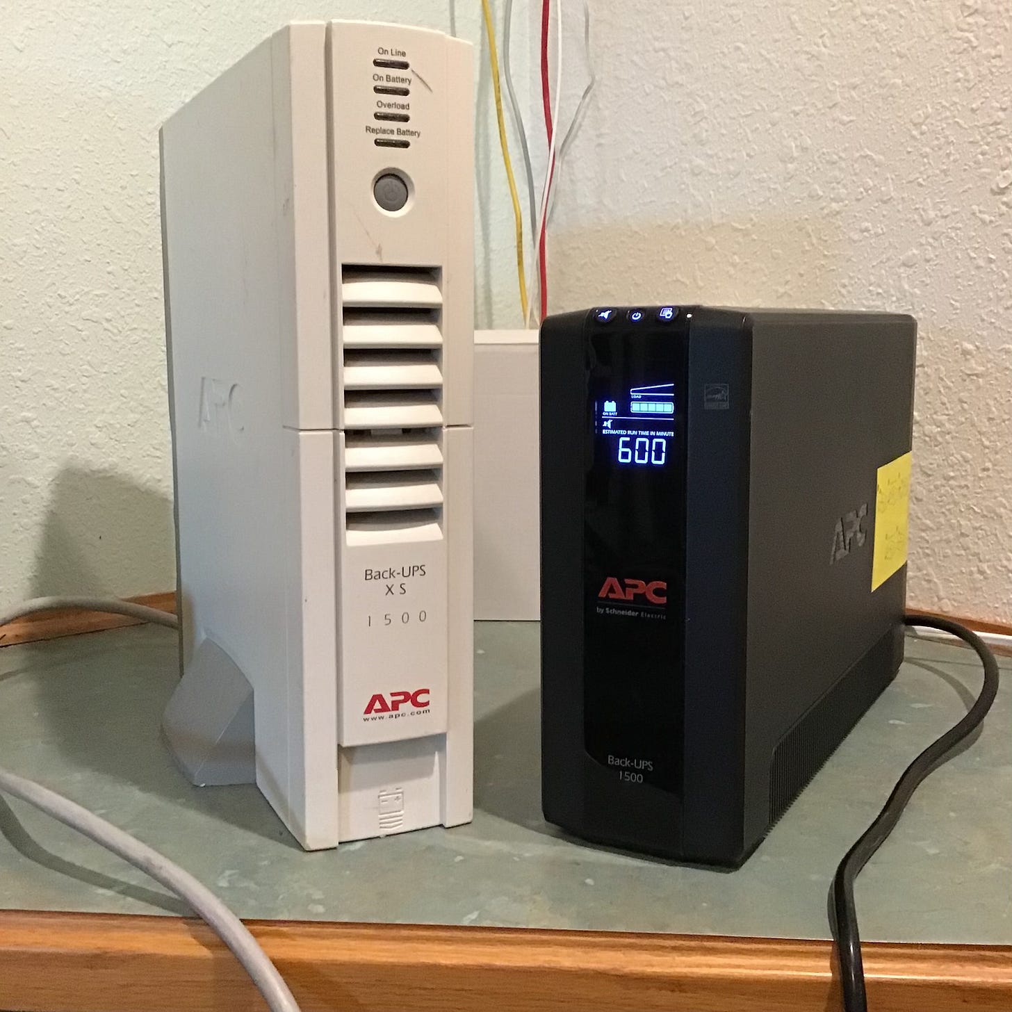 Two APC uninterruptible power supply (UPS) units are side by side on a shelf; the left is an older white Back-UPS XS 1500 and the right is a newer black Back-UPS 1500 displaying “600” on its digital screen.