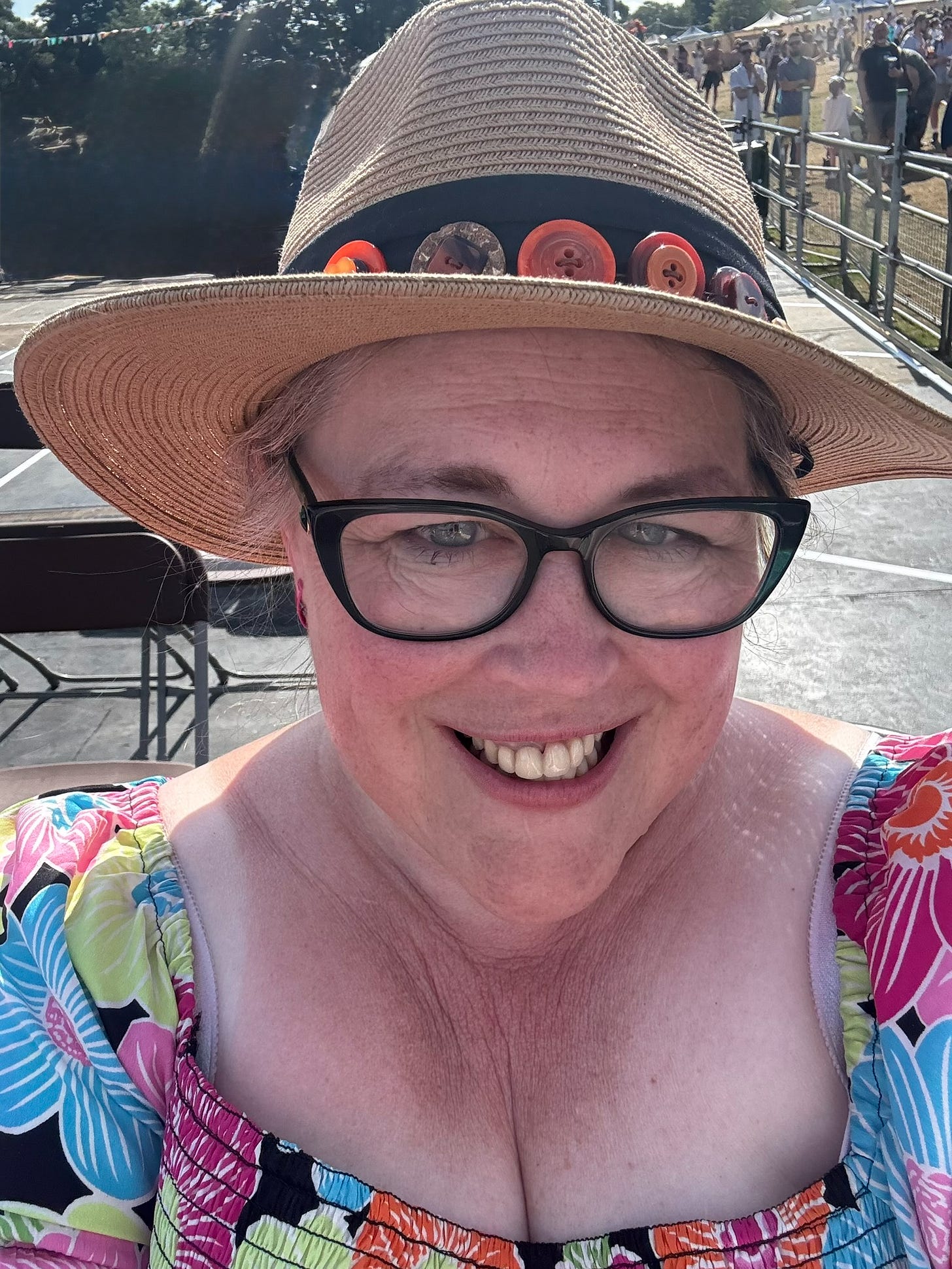 A woman in a straw hat decorated with colourful buttons smiles at an outdoor festival. She is wearing glasses and a bright floral top, with tents and people in the background on a sunny day.