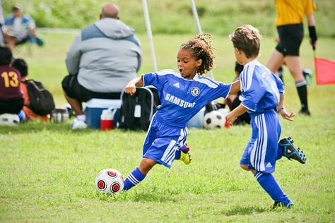 College soccer player in a white Saint Mary’s jersey with number 27 sprints across a sunny field near the goal, with a teammate and the net softly blurred in the background.