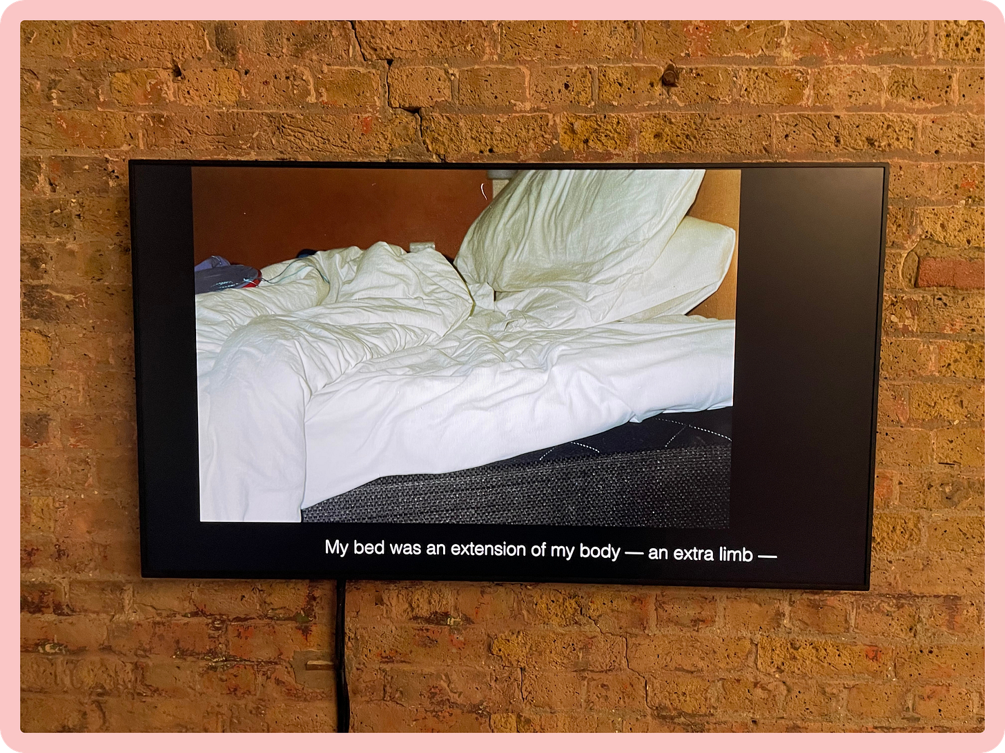 Against a red brick wall, a suspended TV screen shows a photo of an unmade bed, with white sheets and pillows, and below it, a caption which reads "My bed was an extension of my body – an extra limb Against a red brick wall, a suspended TV screen shows a photo of an unmade bed, with white sheets and pillows, and below it, a caption which reads "My bed was an extension of my body – an extra limb