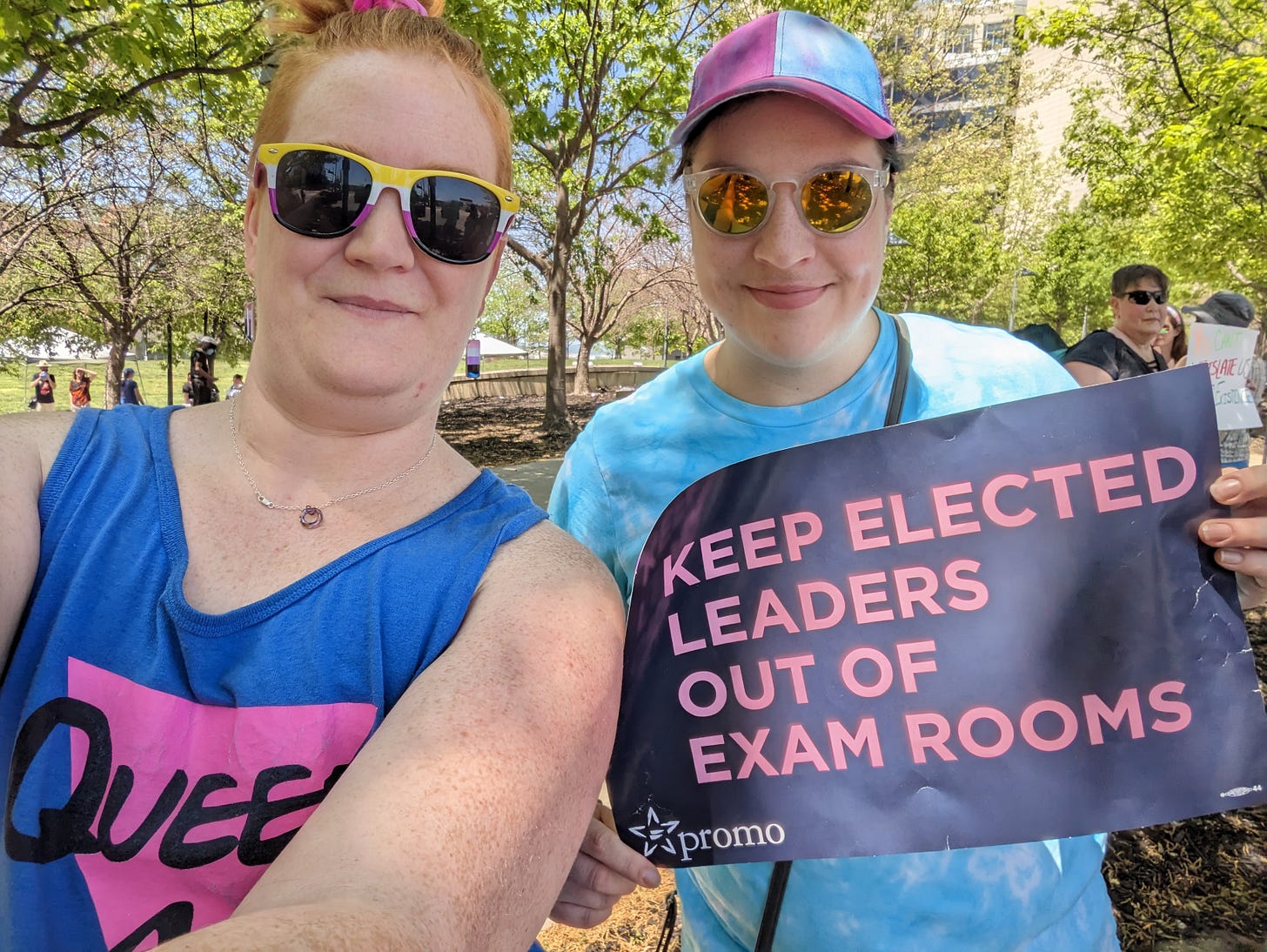 A selfie of two people, a redhead in sunglasses and a brunette in a baseball cap. The first is wearing a tank top that says "Queer AF" and the second is holding a sign that says "KEEP ELECTED LEADERS OUT OF EXAM ROOMS"