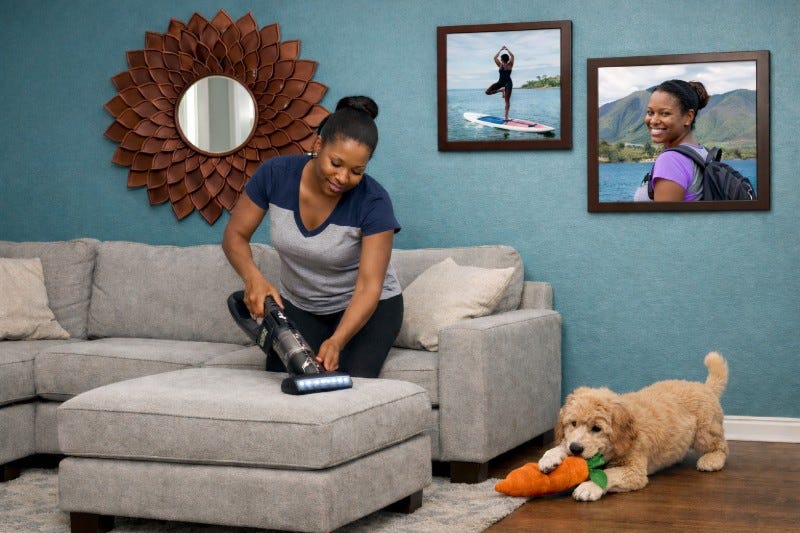 Black woman using a cordless vacuum to clean her ottoman while her Goldendoodle plays with a carrot chew toy. In the background are photos of her in Hawaii and a decorative mirror.