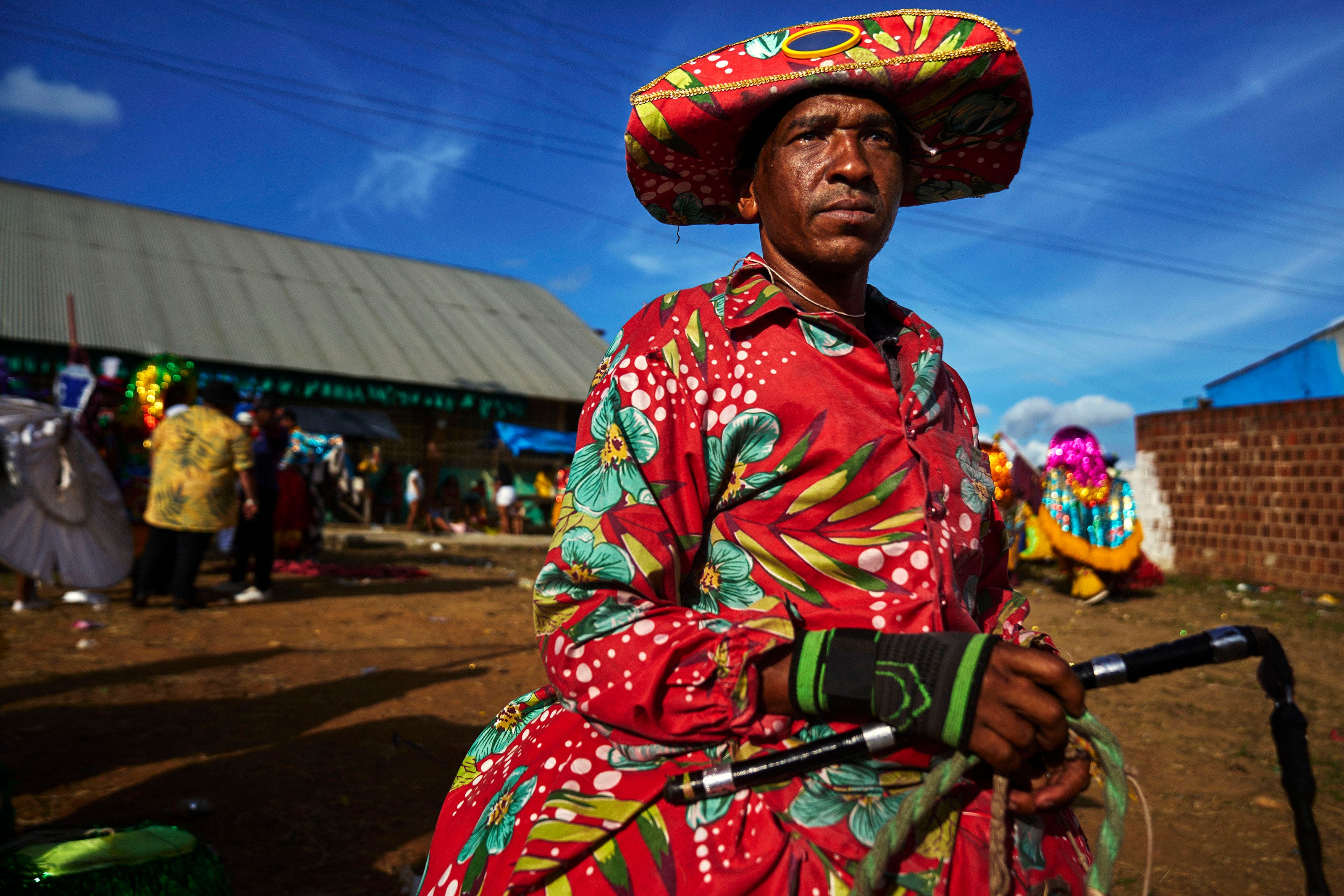 A person wearing a colorful, floral-patterned costume and large matching hat stands outdoors at a festival, holding a cane. Other people in vibrant outfits are visible in the background under a bright blue sky.