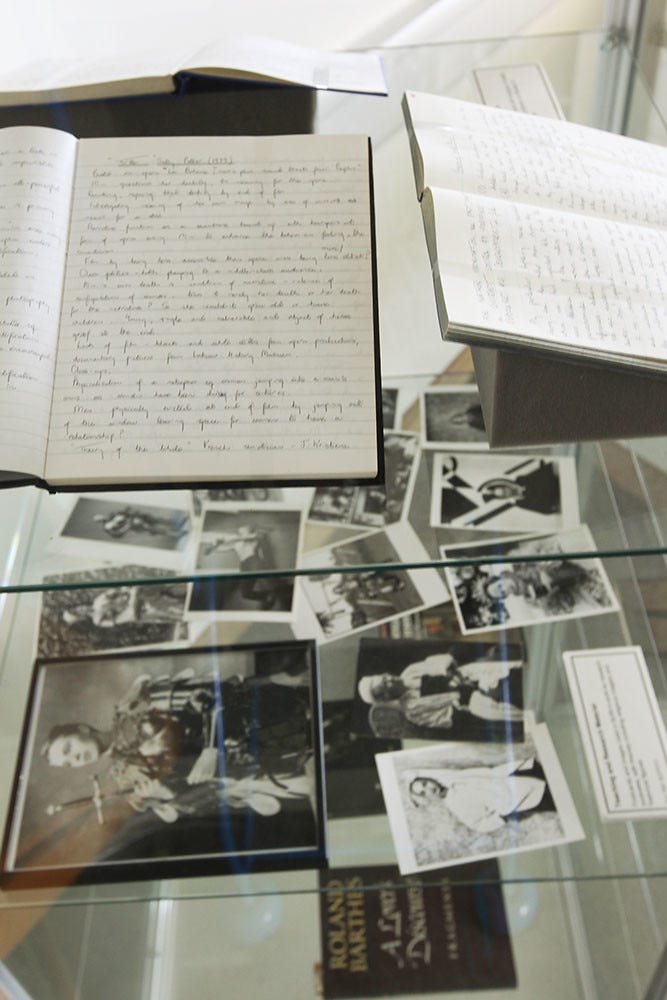 Open notebooks with handwritten text displayed in a glass case alongside vintage black and white photographs, evoking a historical and nostalgic tone. There are also text placards inside that give context to what the viewer is looking at. 