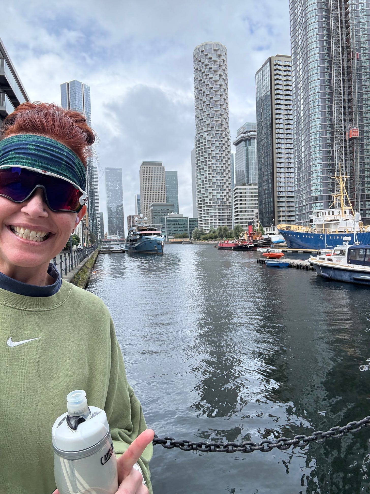 A joyful selfie of Georgina Dean on a walk along the wharf in Canary Wharf, London. She is wearing a green sweatshirt and athletic sunglasses, holding a water bottle. The city skyline and water are visible behind her, capturing a moment of urban exploration.