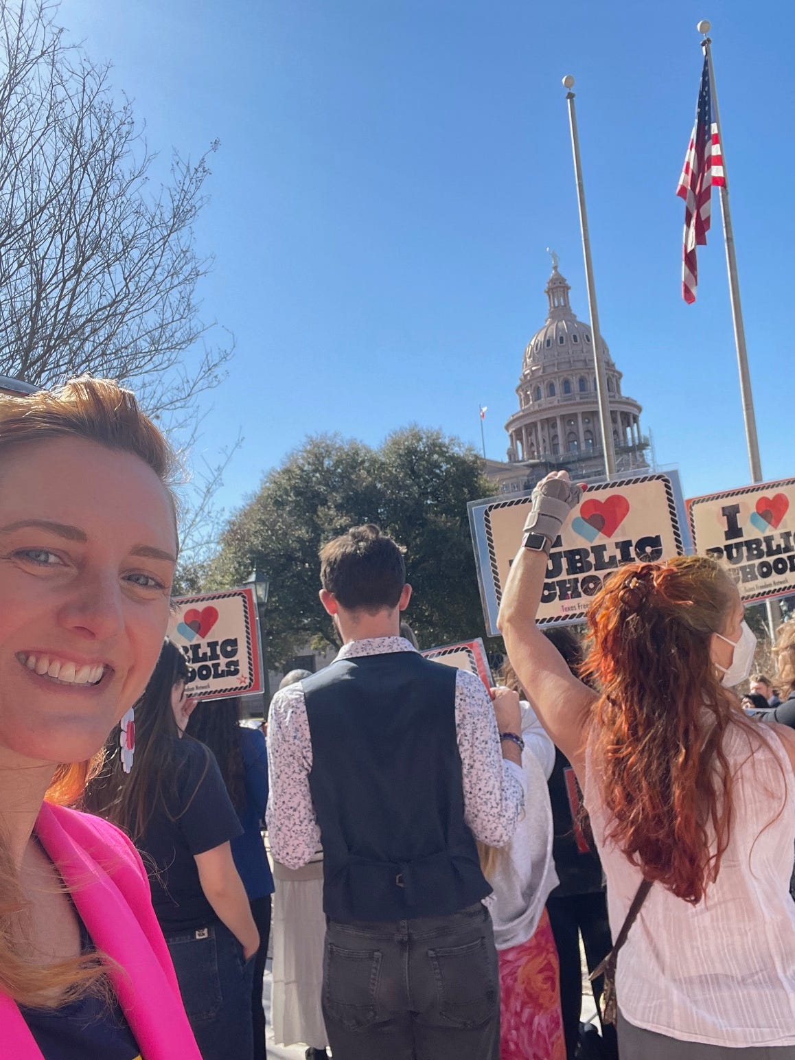 A white woman in a Barbie pink blazer in the foreground smiles at the camera. Behind her many people holding "I love public schools" signs gather in front of the Texas Capitol.