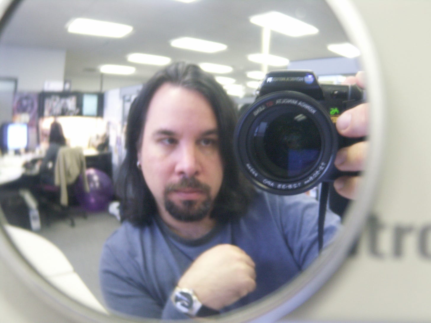 Mirror office selfie of a thirtysomething, long-haired man with a goatee. He's holding up a digital camera to a fish-eye rearview mirror stuck to his computer monitor