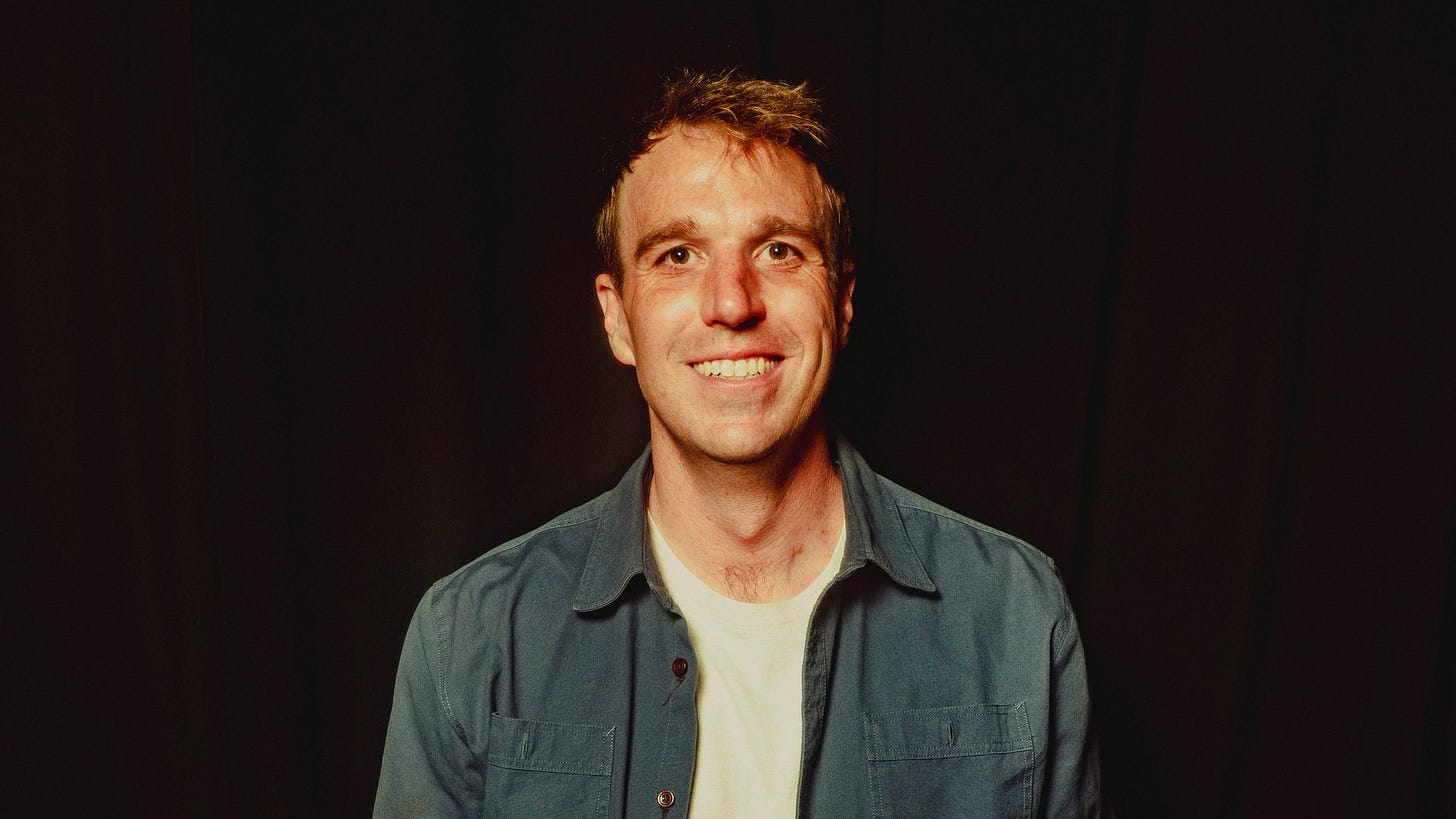 Luke Strathmann smiling in a denim shirt under warm stage light.