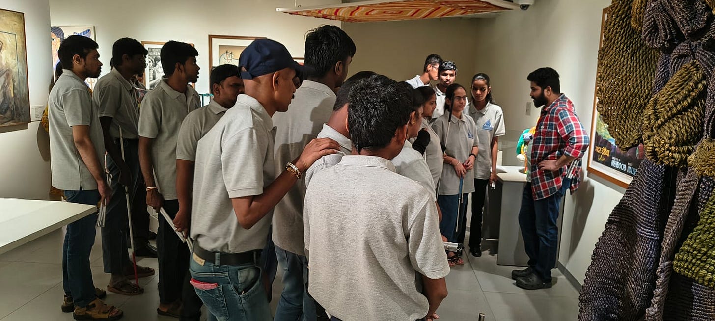 Shailesh is dressed in a white and blue short and black jeans. He is talking to a group of blind students at an exhibition at MAP. The walls behind them are filled with paintings, mostly hidden in the photo. Shailesh is dressed in a white and blue short and black jeans. He is talking to a group of blind students at an exhibition at MAP. The walls behind them are filled with paintings, mostly hidden in the photo.