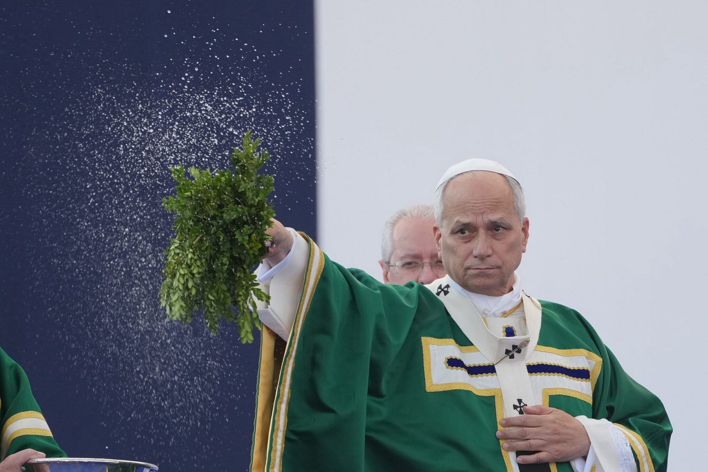 Pope Leo XIV, in green vestments, sprinkles holy water over the congregation with a sprig of greenery at an outdoor Mass. Pope Leo XIV, in green vestments, sprinkles holy water over the congregation with a sprig of greenery at an outdoor Mass.