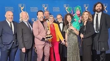 A group of men and women smile towards the camera with one holding an Emmy Award and one holding a puppet of Mokey Fraggle.