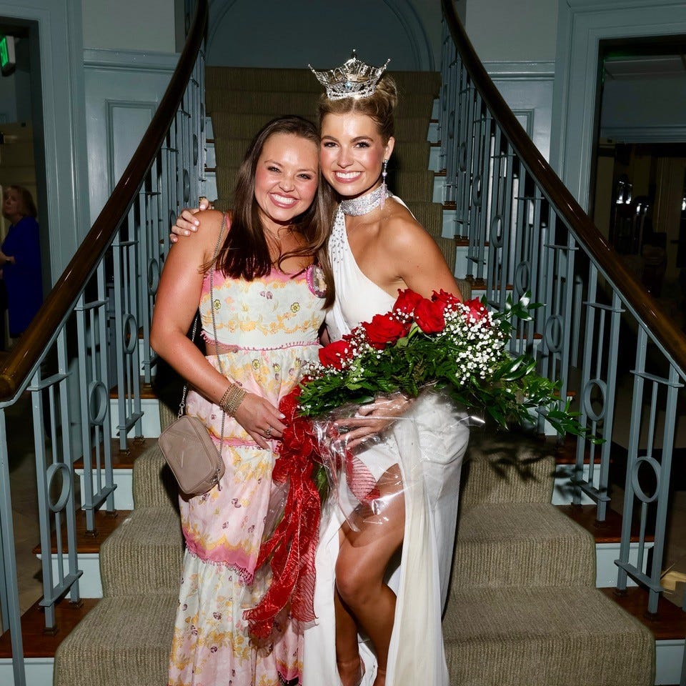 A light-skinned woman wearing a floral dress stands next to a light-skinned woman wearing a white ball gown, a crown, and holding a bouquet of red roses in her arms.