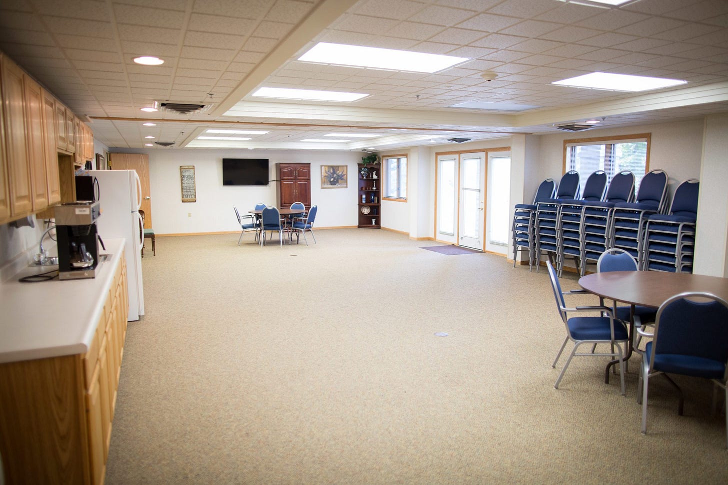 A white rectangular room in a nursing home, lit by fluorescent lights, with two tables and dozens of chairs stacked along one wall. A white rectangular room in a nursing home, lit by fluorescent lights, with two tables and dozens of chairs stacked along one wall.