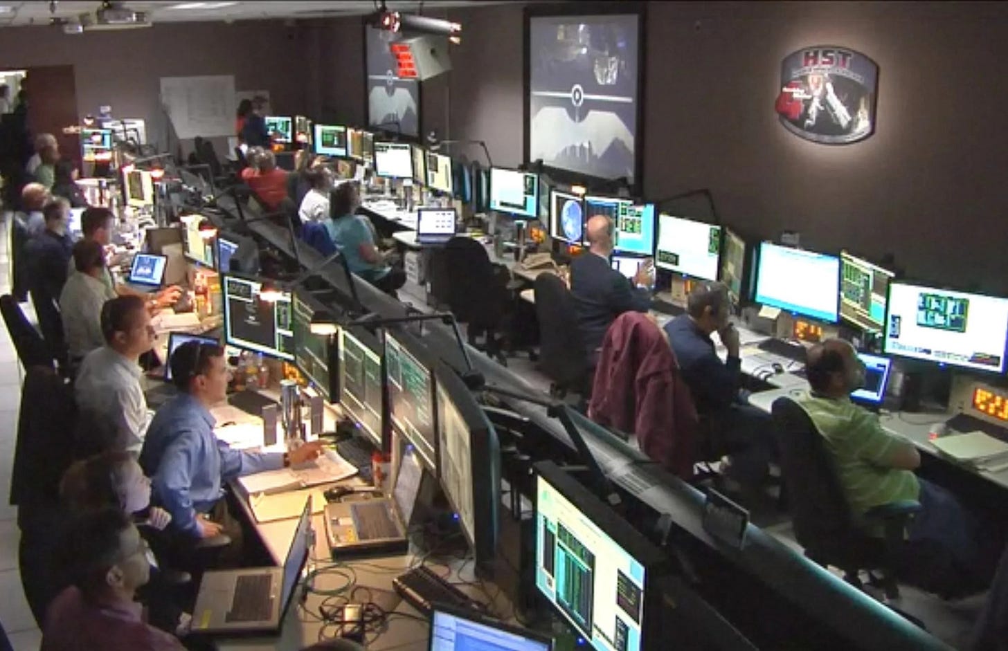Photograph of a group of engineers sitting in front of their computers inside of the "Space Telescope Operations Control Center." This image was taken during Hubble's Servicing Mission 4, which is on the screen in the front of the room. Photograph of a group of engineers sitting in front of their computers inside of the "Space Telescope Operations Control Center." This image was taken during Hubble's Servicing Mission 4, which is on the screen in the front of the room.