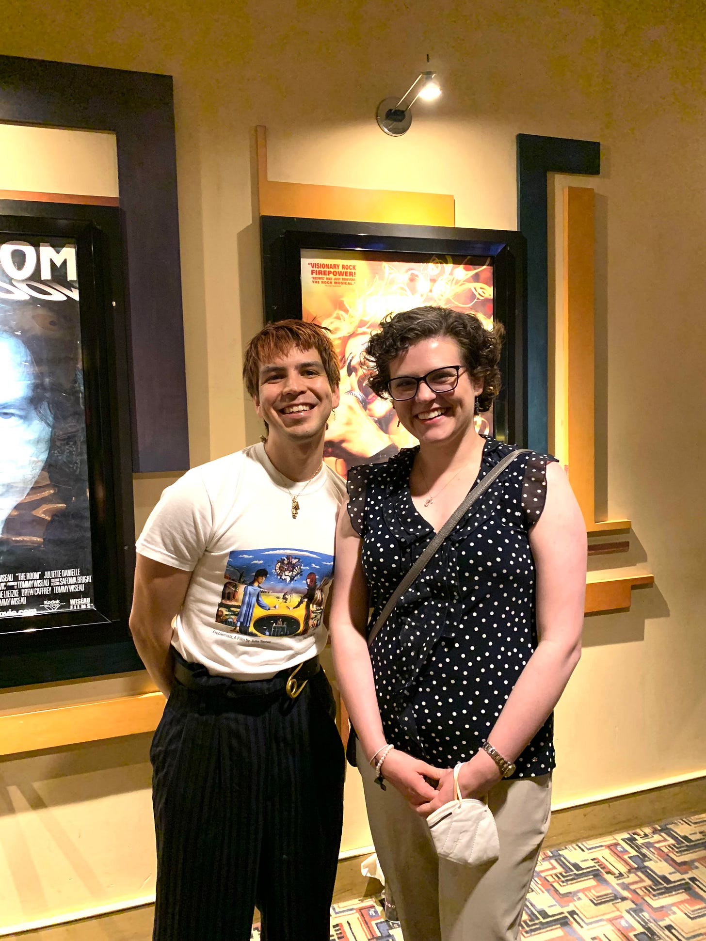 Julio Torres and Emily. Julio is wearing a problemista t-shirt and a gold chain. His hair is bleached. They are both smiling big.