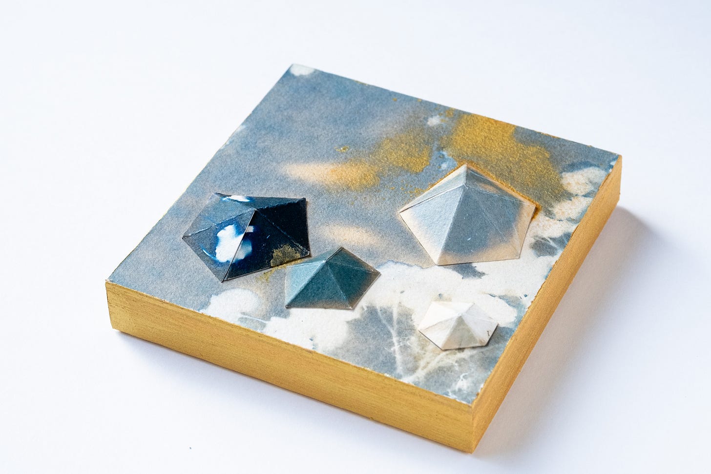 The edges of each panel are painted with gold acrylic, in addition to the surface of the light blue (toned) cyanotype being sprinkled with gold acrylic. The geometric bumps are different shades than the underlying surface.
