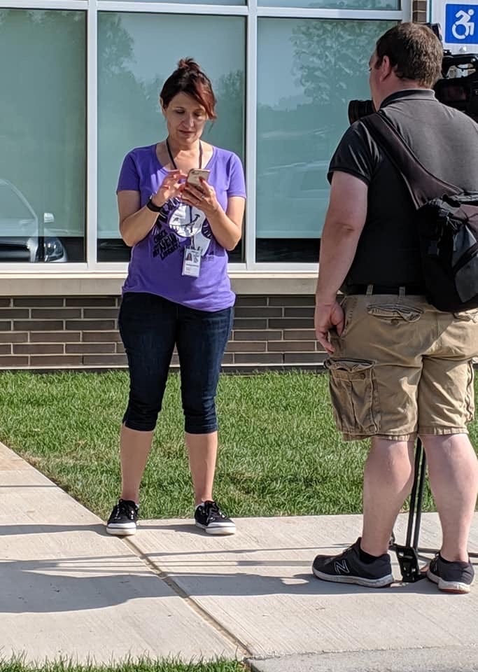 Photo of me, a white lady with brown hair wearing jeans and a purple t-shirt, absorbed in something on my phone while a reporter with a film camera waits patiently for me to pay attention to him. I was super-young here. Holy cow. Photo of me, a white lady with brown hair wearing jeans and a purple t-shirt, absorbed in something on my phone while a reporter with a film camera waits patiently for me to pay attention to him. I was super-young here. Holy cow.