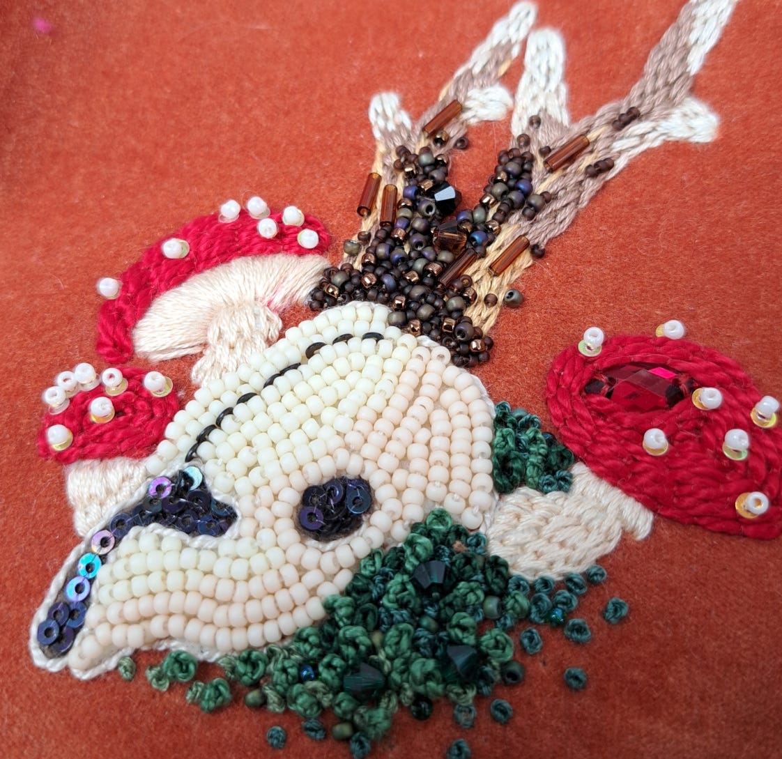 A hand embroidered deer skull motif, featuring fly agaric and moss, using various beads and threads