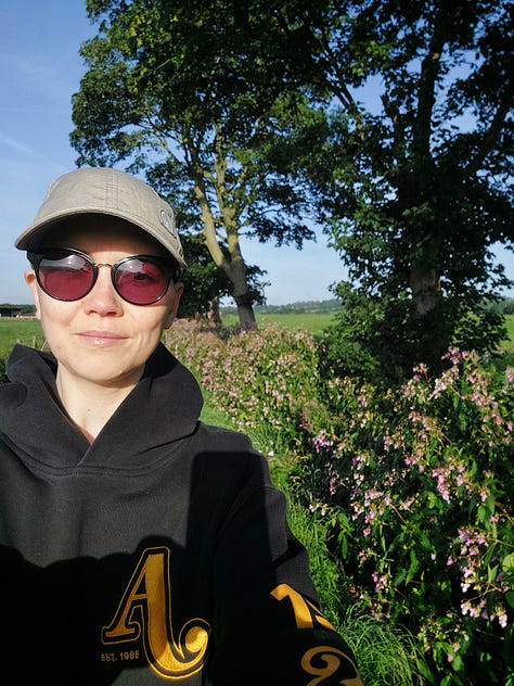 Smiling selfies of me mid walk. Behind me you see pink blooming Himalayan Balsam. In another picture is a straight grey road disappearing into the horizon. In another is a body of water, some grey clouds and lush grass in the foreground. Other pictures are of nature, a close up of an orange and black painted lady butterfly, dappled sun shining through woodland pines and a canal with reeds along its edge and algae on the water