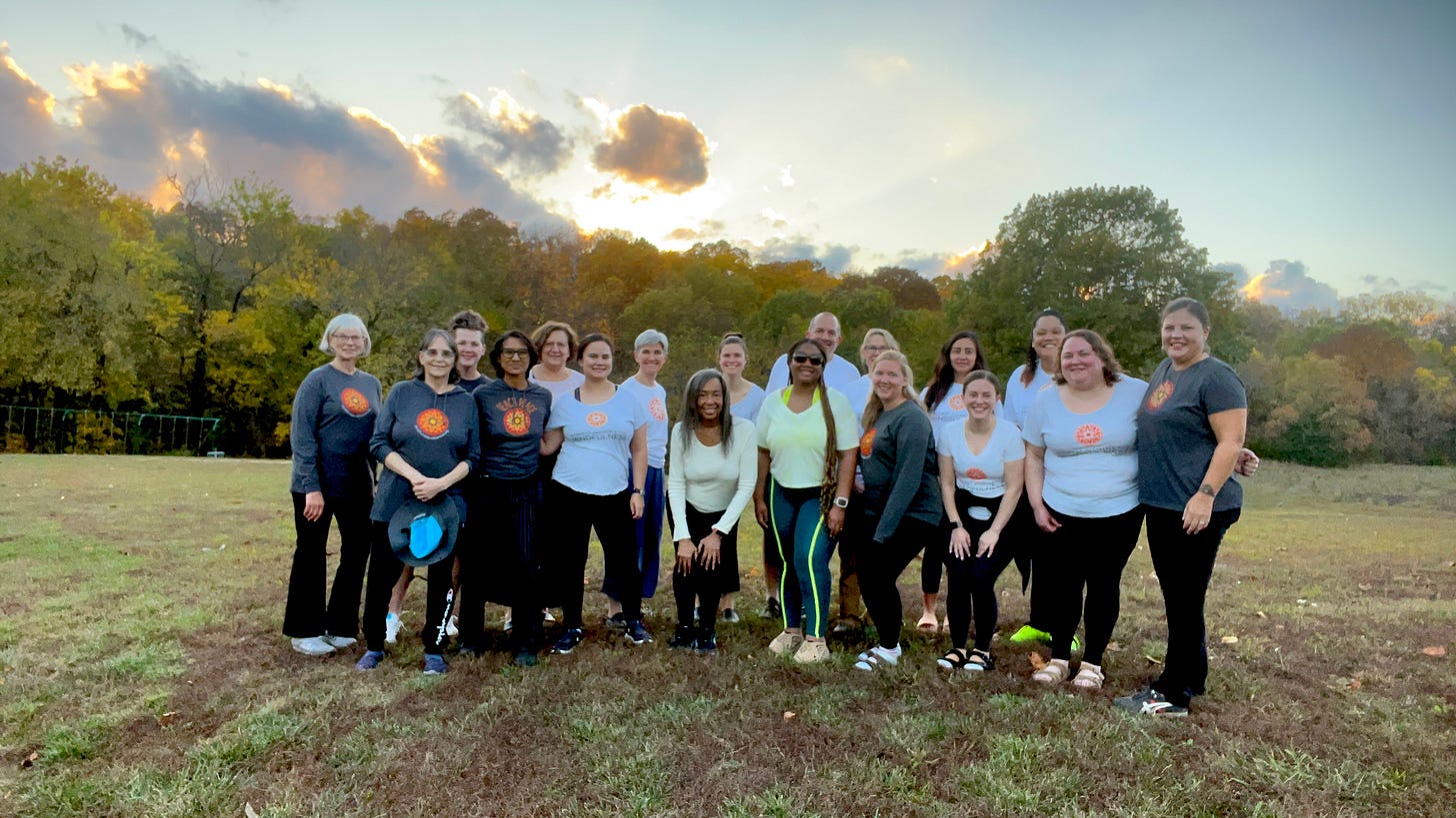 a group of people standing on a grassy hill against a backdrop of trees with fall colored leaves and a colorful sunset