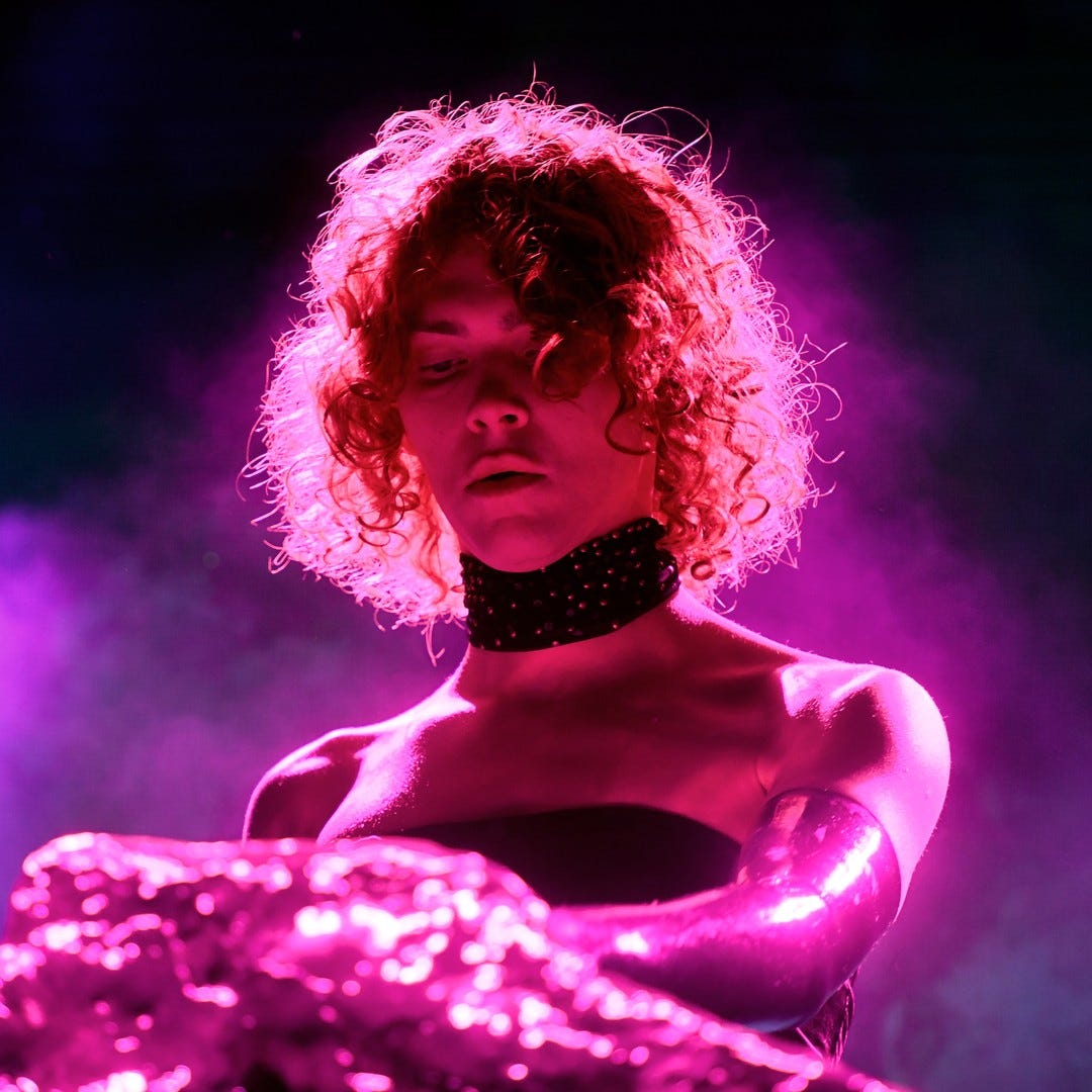A photograph of a redhead wearing a bejeweled choker, swathed in neon lights and fog.