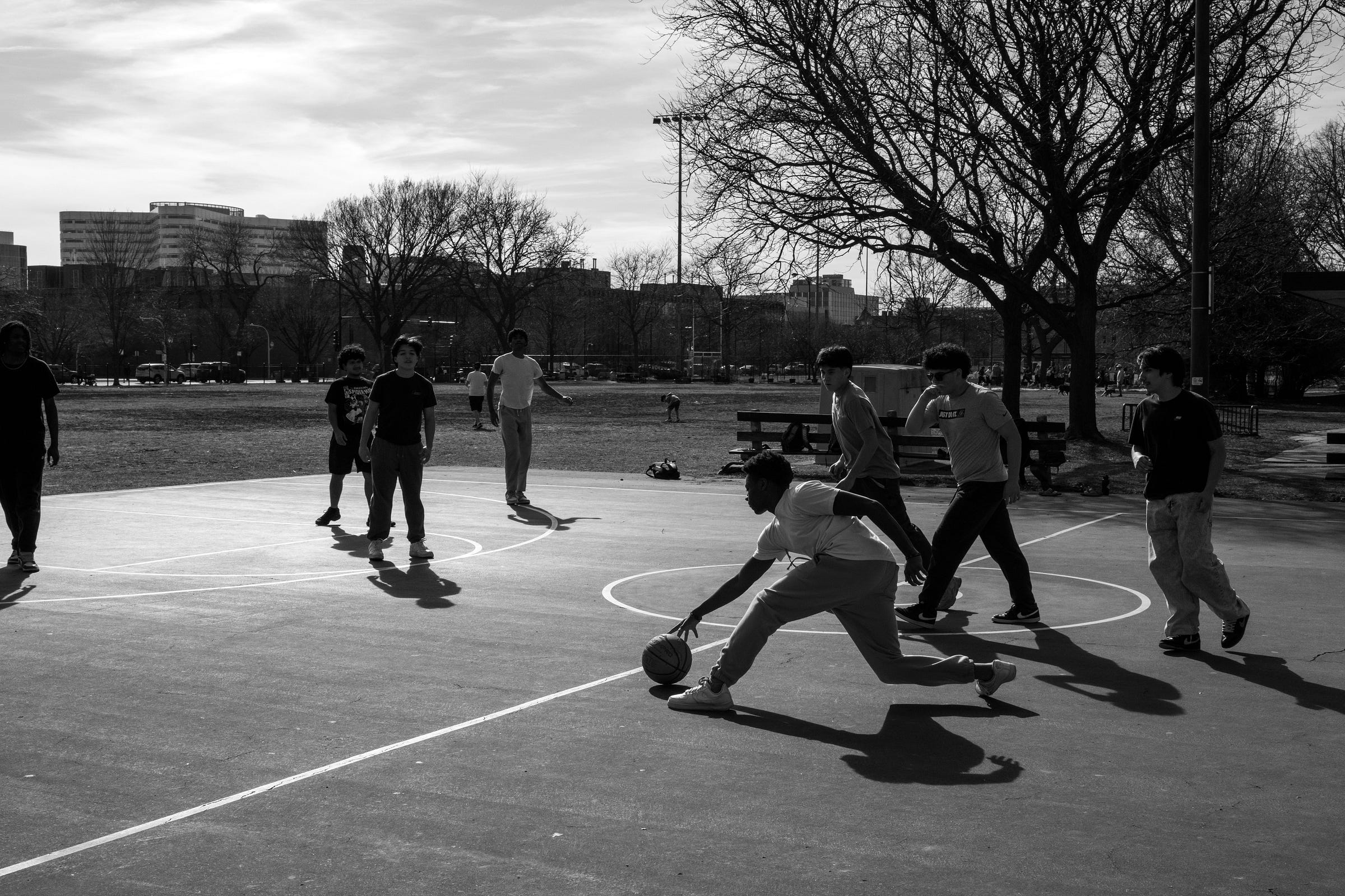 This is a powerful black-and-white photograph capturing a group of young people playing basketball on an outdoor court. The contrast between the shadows and the highlights adds depth and emotion to the scene, emphasizing movement and connection within the community.