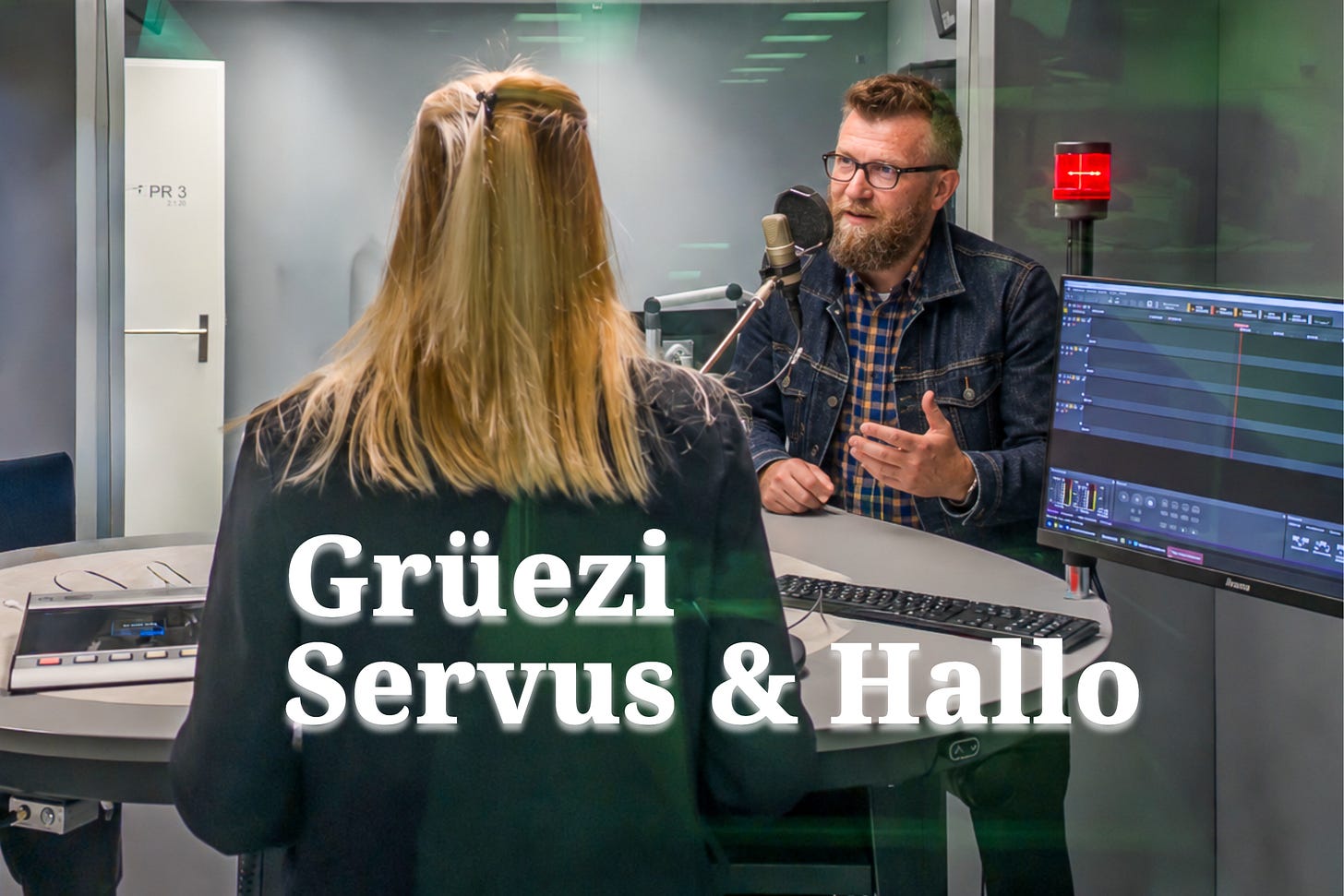 Interview im Radiostudio: Jürg Vollmer mit Bart und Brille steht einer blonden Rediojournalistin gegenüber und spricht gestikulierend in ein Mikrofon. Rechts ist die Audiotechnik zu sehen. Darunterder Schriftzug «Grüezi Servus & Hallo».