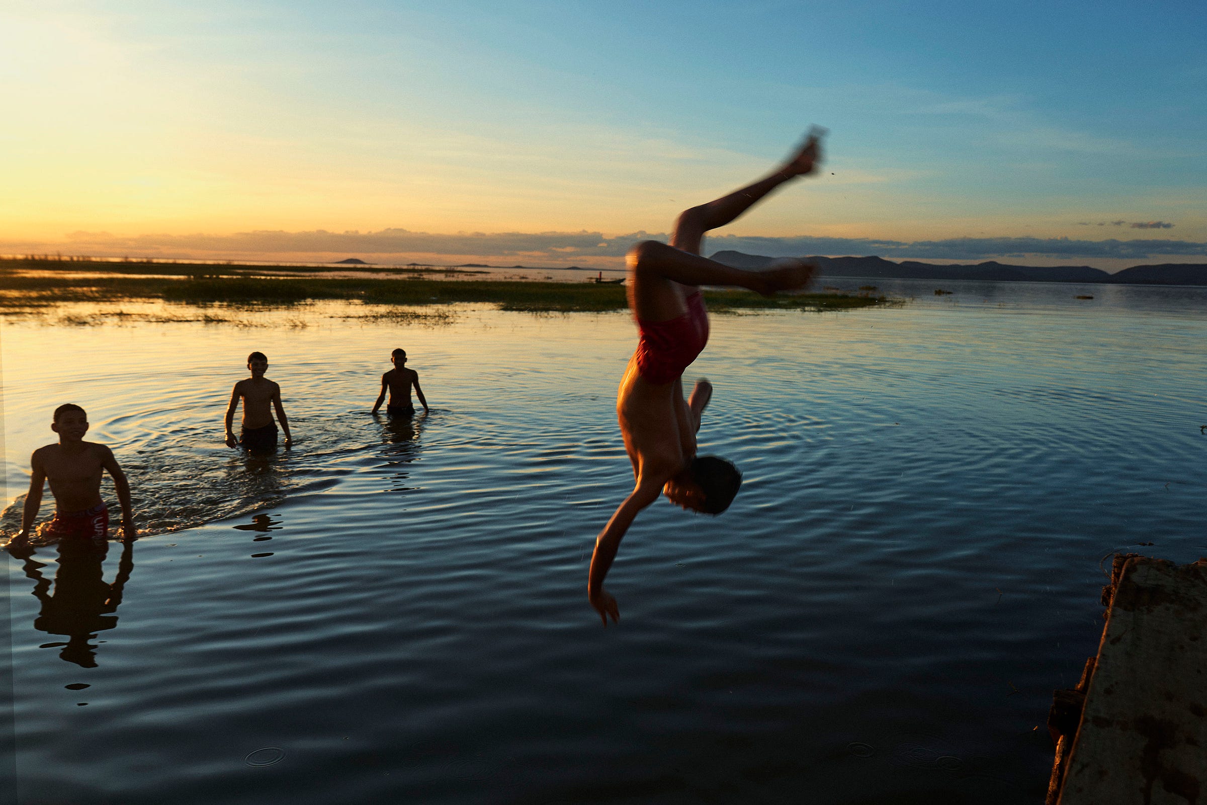 A child mid-flip dives into a calm lake at sunset, while three other children stand waist-deep in the water, silhouetted against the colorful sky.