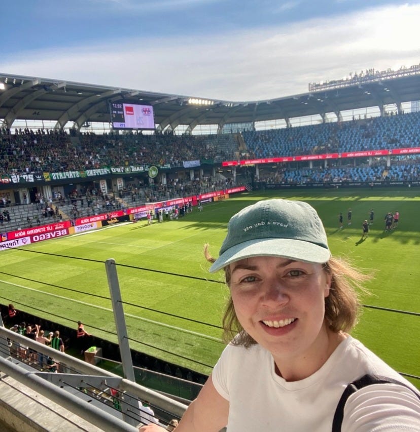 Woman in a green cap taking a selfie at a football stadium, with players warming up on the pitch and a full stand of fans behind her. Woman in a green cap taking a selfie at a football stadium, with players warming up on the pitch and a full stand of fans behind her.