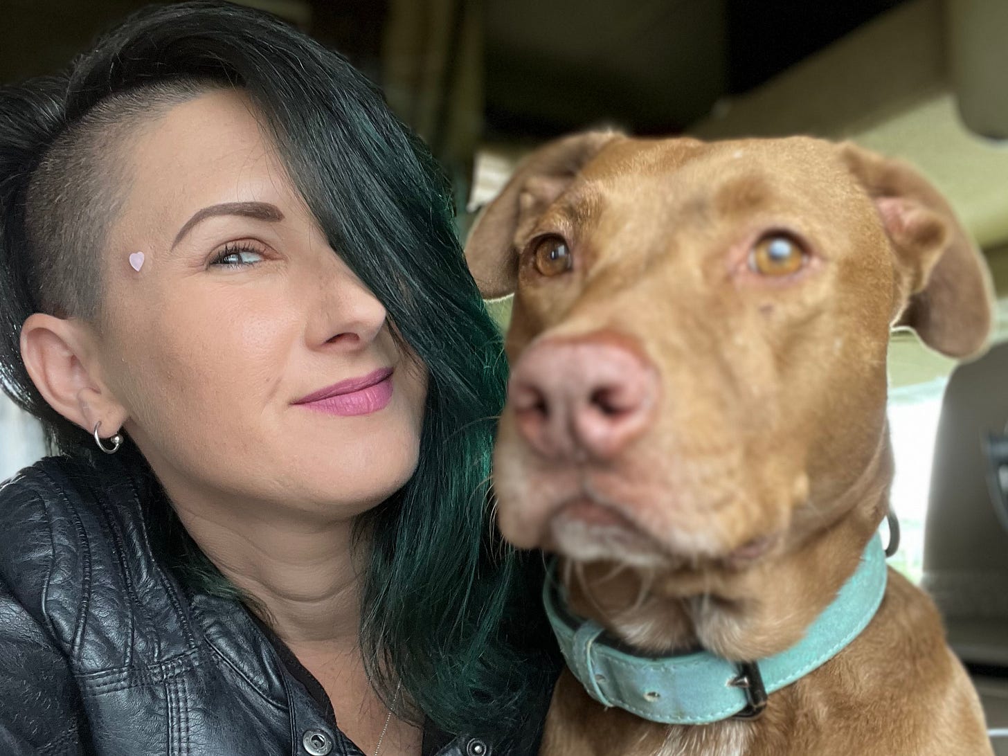 Lyric, with long green and black hair, wearing a black leather jacket and posing with their friend Sadie, the pit bull, who is reddish brown in color and wearing a teal collar. Lyric, with long green and black hair, wearing a black leather jacket and posing with their friend Sadie, the pit bull, who is reddish brown in color and wearing a teal collar.