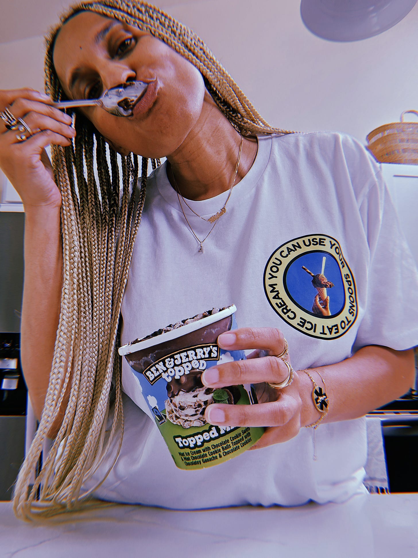Dr. Cadet standing in her kitchen eating from a pint of Ben & Jerry’s ice cream wearing her new t-shirt. The white t-shirt has a picture of an ice cream cone with chocolate ice cream, adorned by a waffle straw swirled with chocolate and a neon yellow spoon set in a electric blue and green circle that gives 90s vibes. The ice cream cone is surrounded by the words “you can use your spoon for ice cream” and is held by a Black disabled woman who chose using one of her spoons for joy (and you should too). Photographer: Dr. Akilah Cadet