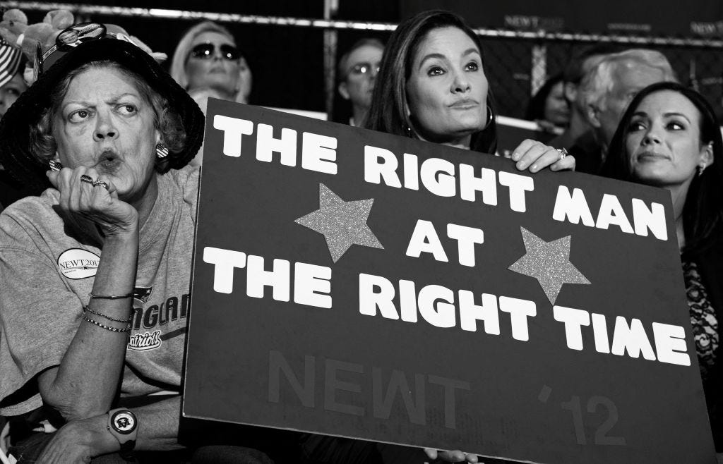 Three women sit in a crowd, one holding a large sign that reads “THE RIGHT MAN AT THE RIGHT TIME” with “NEWT ‘12” partially visible at the bottom.