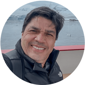 20260810_Marcos-Santos_IOUSP Homem no convés de uma embarcação. Ele tem cabelos curtos e escuros, veste um casaco acolchoado preto para frio intenso e usa uma mochila com alça visível sobre o ombro esquerdo. Ao fundo, uma paisagem polar. O mar está calmo, com pequenos blocos de gelo flutuando sobre a superfície